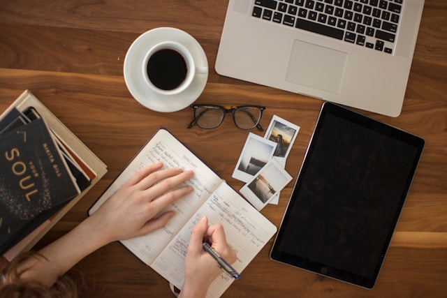 Overhead view of a wooden desk with a person writing in an open notebook beside a cup of coffee, laptop, tablet, glasses, books, and small printed photos.